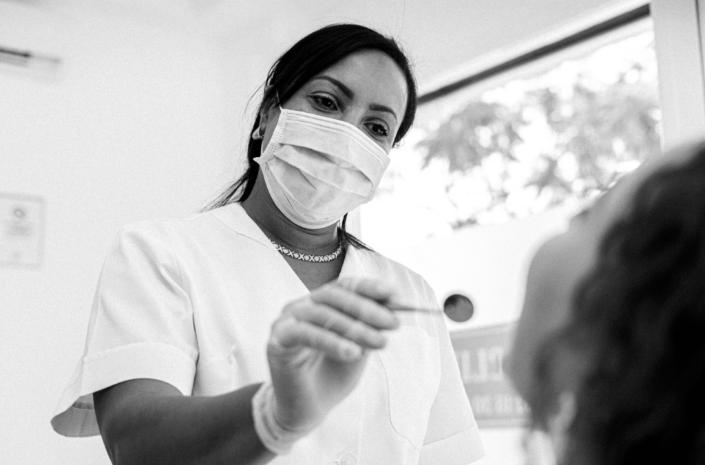 Fotografía en blanco y negro de una dentista usando mascarilla y guantes, sosteniendo un instrumento mientras atiende a un paciente.