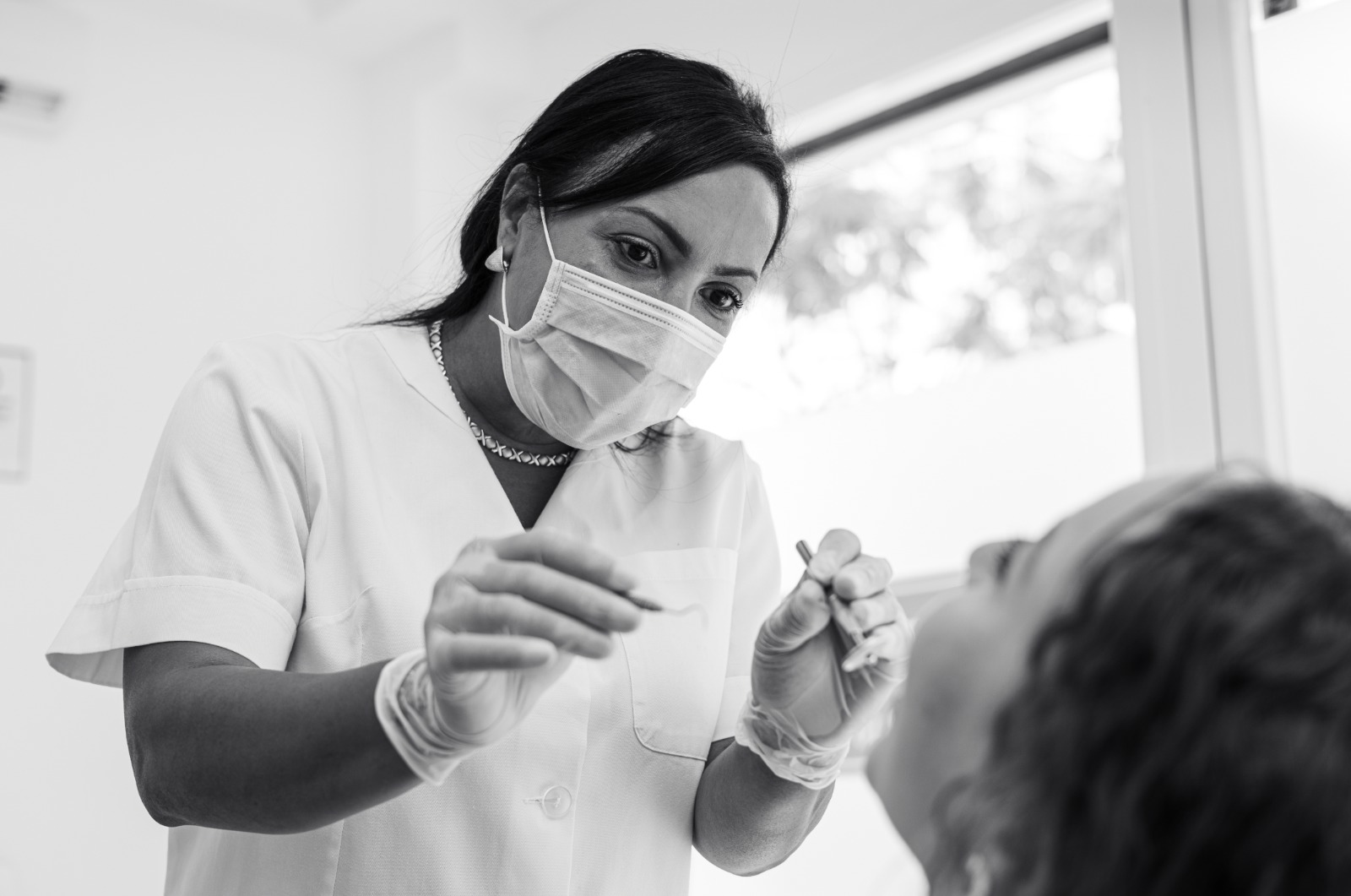 Fotografía en blanco y negro de una dentista, con mascarilla y guantes, examinando la boca de un paciente usando un instrumento dental.