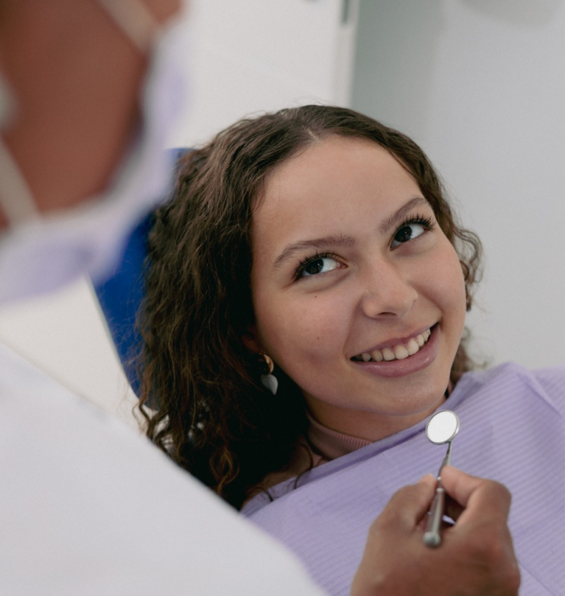 Fotografía a color de una paciente sonriendo mientras la dentista sostiene un espejo dental cerca de su boca.